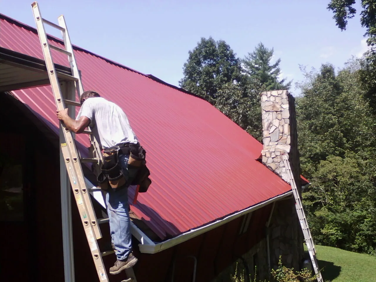 Roofer on a ladder beside a red metal roof and stone chimney — Soffit & Fascia Repair by Roofing Brothers in Canandaigua